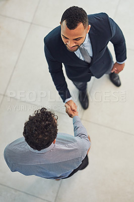 Buy stock photo High angle shot of two confident young businessmen shaking hands in agreement while standing in the office during the day