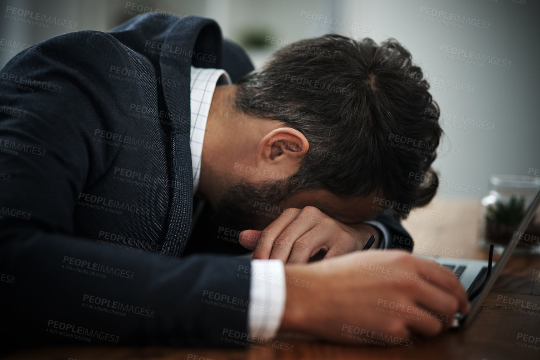 Buy stock photo Shot of a young businessman lying with his head down on a laptop in an office