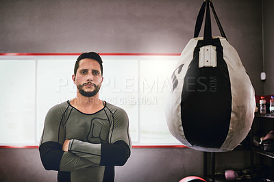 Buy stock photo Portrait of a confident young male boxer standing with his arms folded next to a punching bag inside of a gym during the day