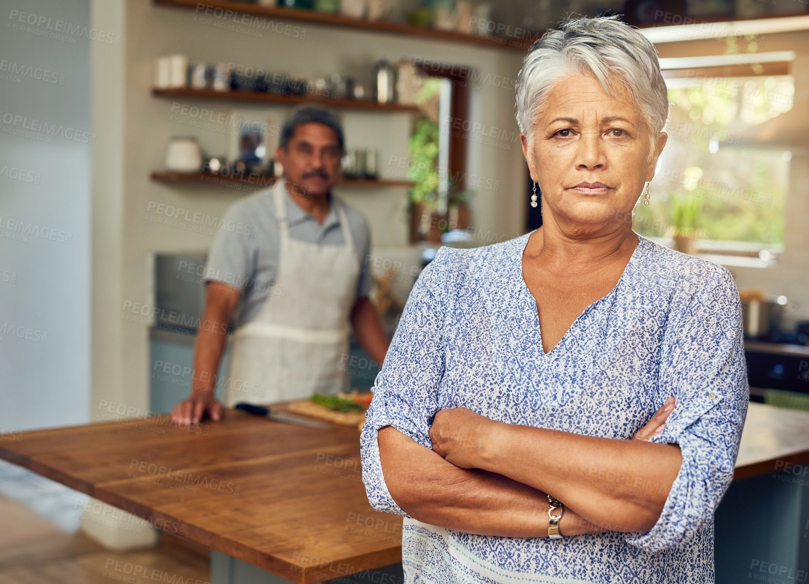 Buy stock photo Portrait of a mature woman looking upset after a fight with her husband who is cooking in the background