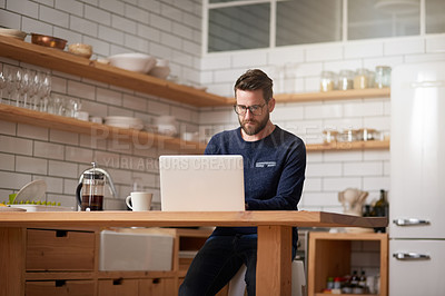 Buy stock photo Cropped shot of a handsome young businessman working on his laptop while sitting in the kitchen at home