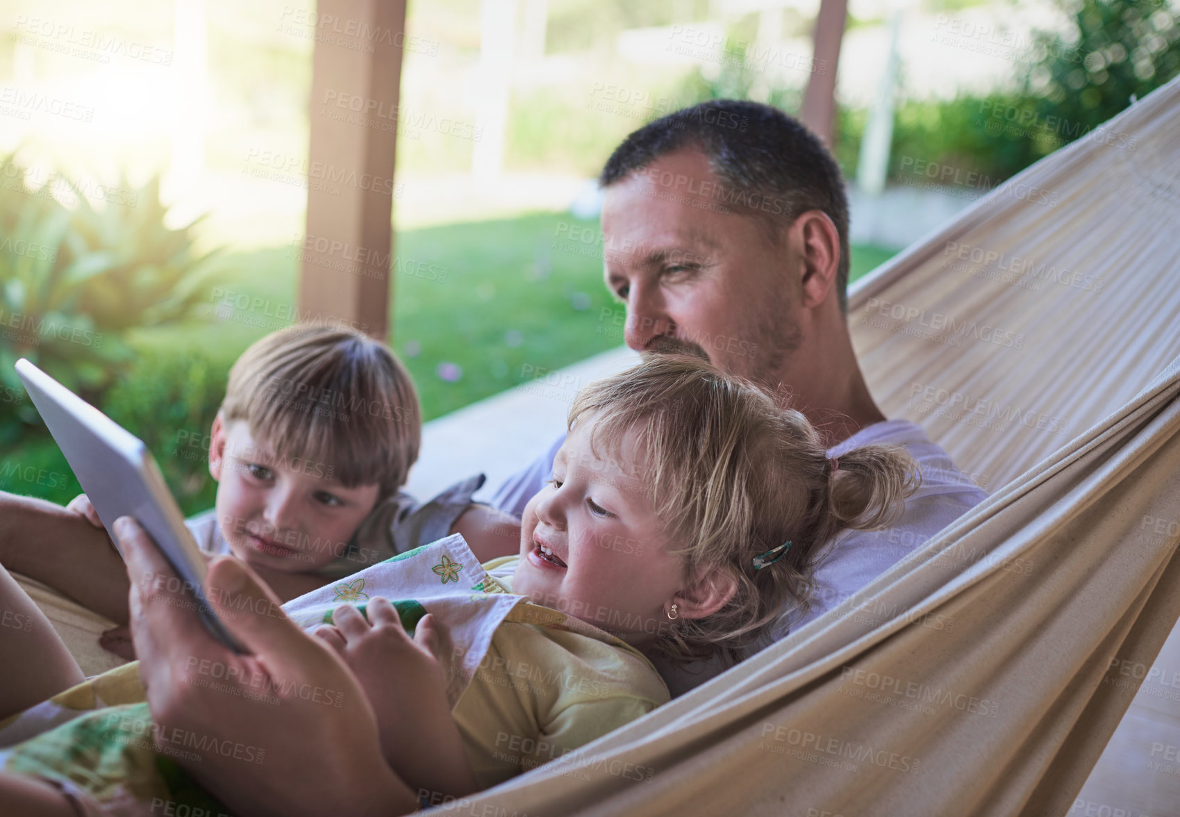 Buy stock photo Shot of a father and his two little children using a digital tablet while relaxing on a hammock outdoors