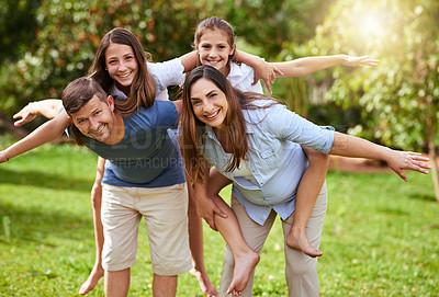 Buy stock photo Portrait of a cheerful young mother and father giving their daughters a piggyback ride outside in a park during the day