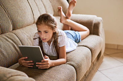 Buy stock photo Shot of a focussed young girl browsing on a digital tablet while lying on a sofa at home during the day