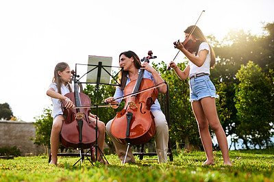 Buy stock photo Shot of a cheerful young mother and her two daughters playing classical string instruments together while standing in the backyard of their home