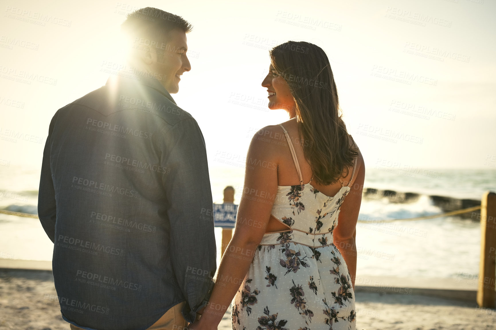 Buy stock photo Rearview shot of an affectionate young couple holding hands while standing face to face on the beach