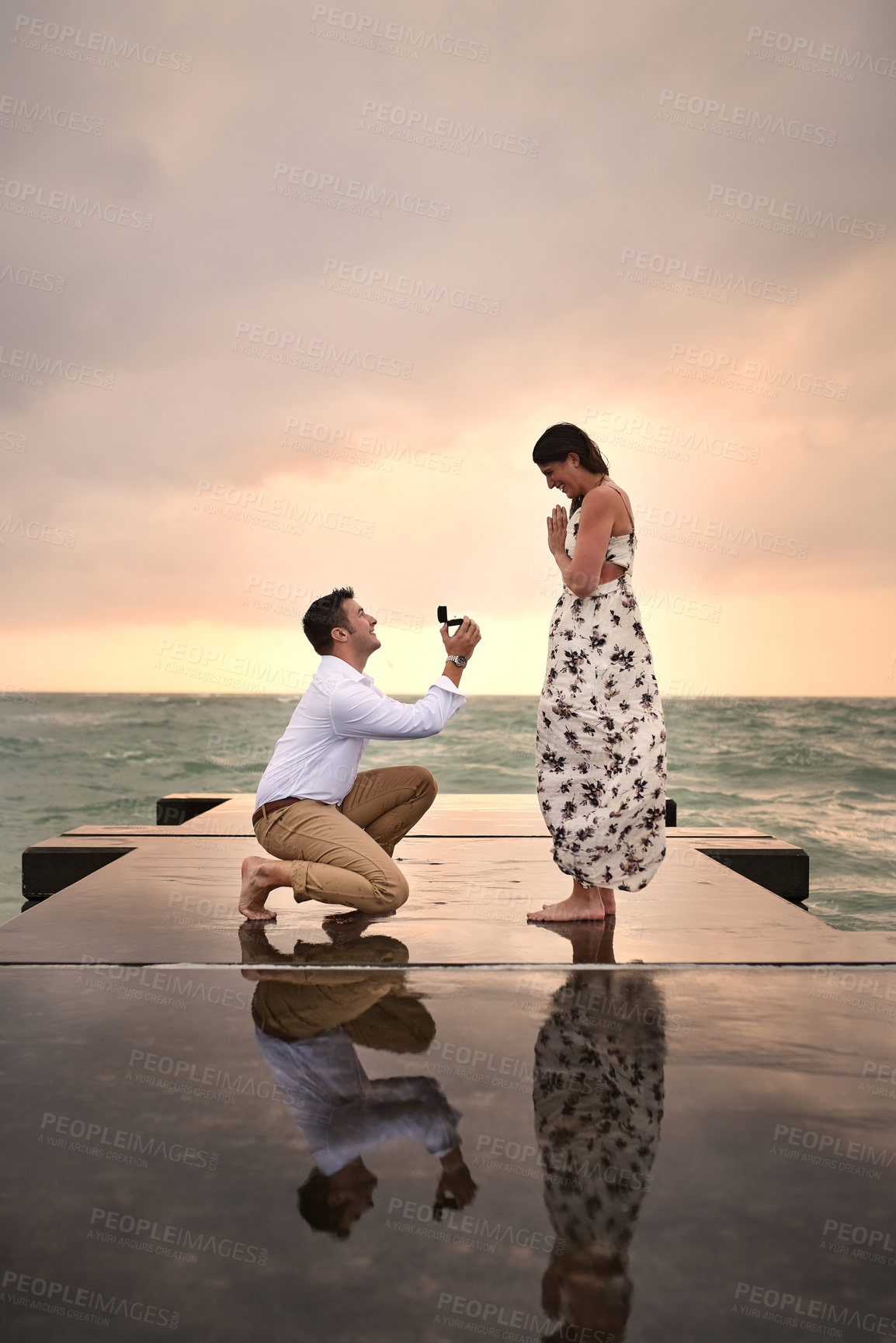 Buy stock photo Full length shot of a handsome young man proposing to his girlfriend on the jetty at the beach