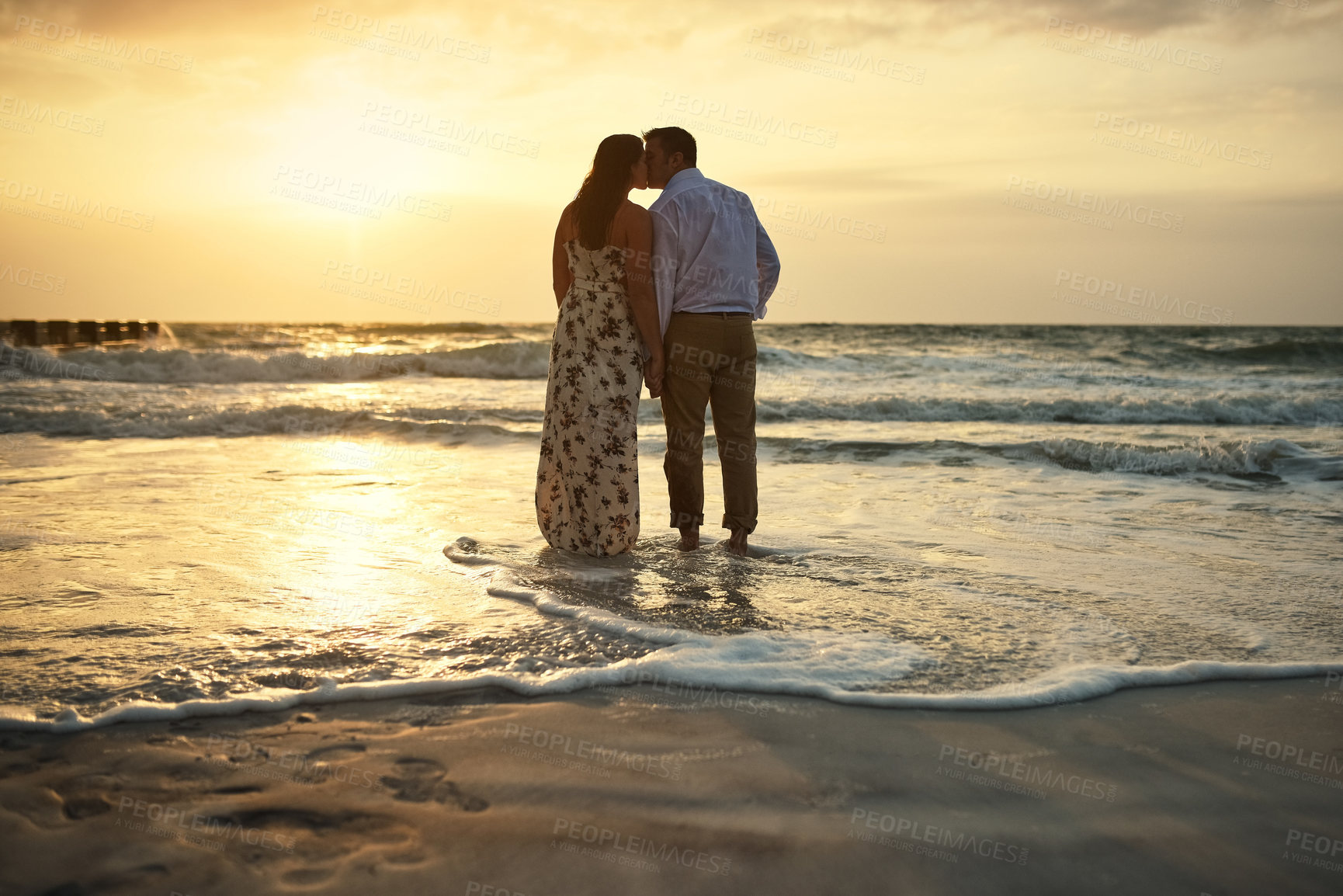 Buy stock photo Rearview shot of an affectionate young couple holding hands while kissing on the beach
