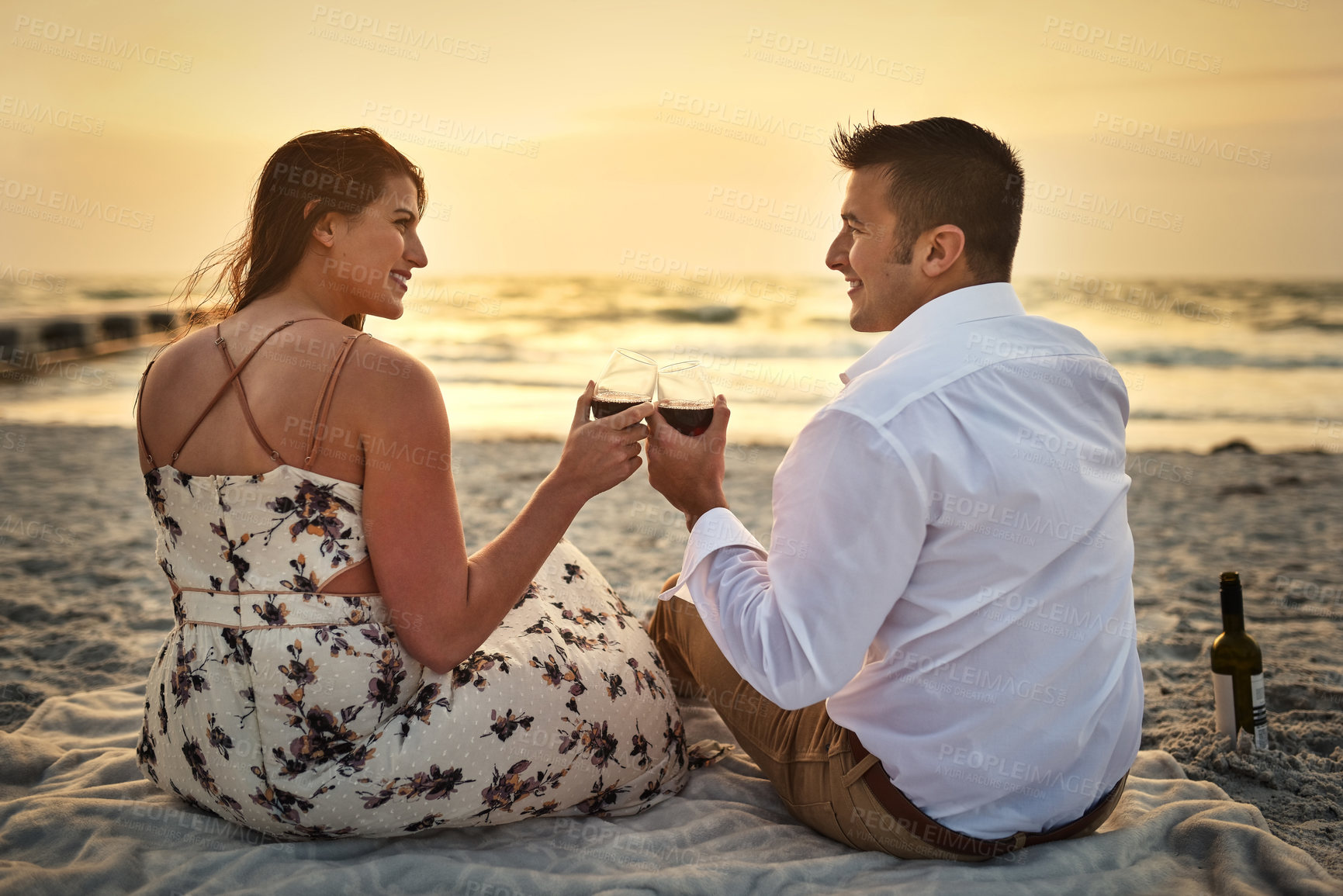Buy stock photo Rearview shot of an affectionate young couple enjoying some wine while sitting on the beach