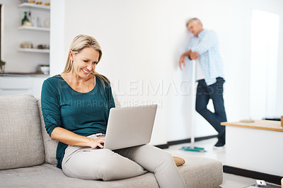 Buy stock photo Shot of a mature woman using a laptop at home while her husband sweeps in the background