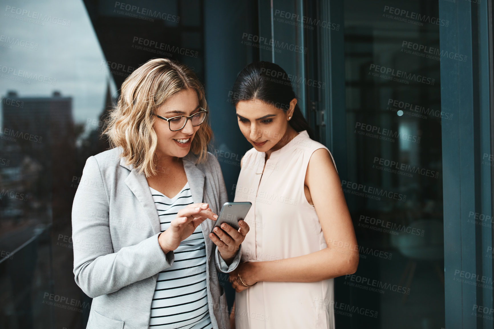 Buy stock photo Shot of two businesswomen using a cellphone together on the office balcony