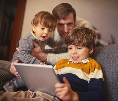 Buy stock photo Shot of an adorable little boy using a tablet on the sofa at home while his father and younger brother look on