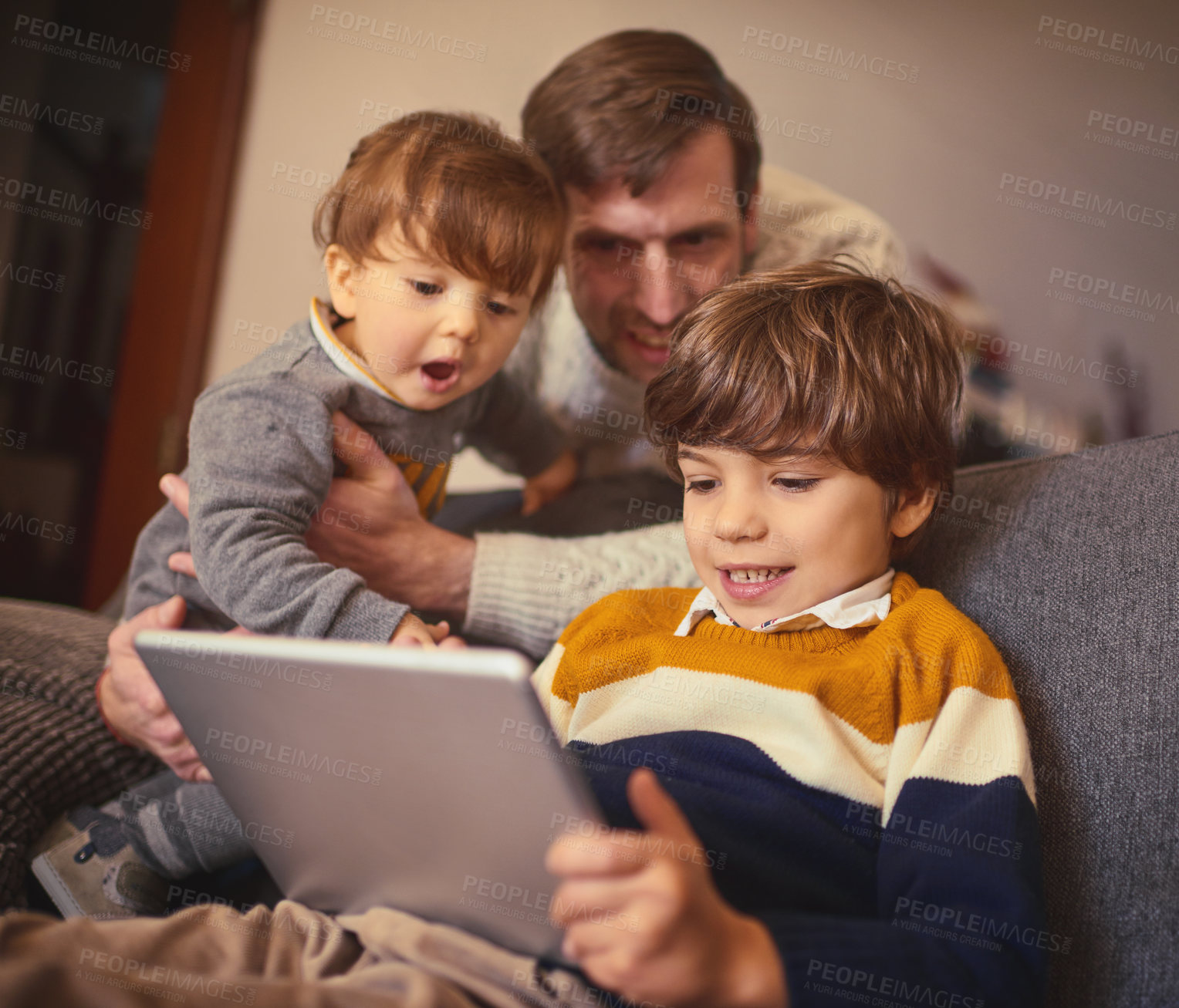 Buy stock photo Shot of an adorable little boy using a tablet on the sofa at home while his father and younger brother look on
