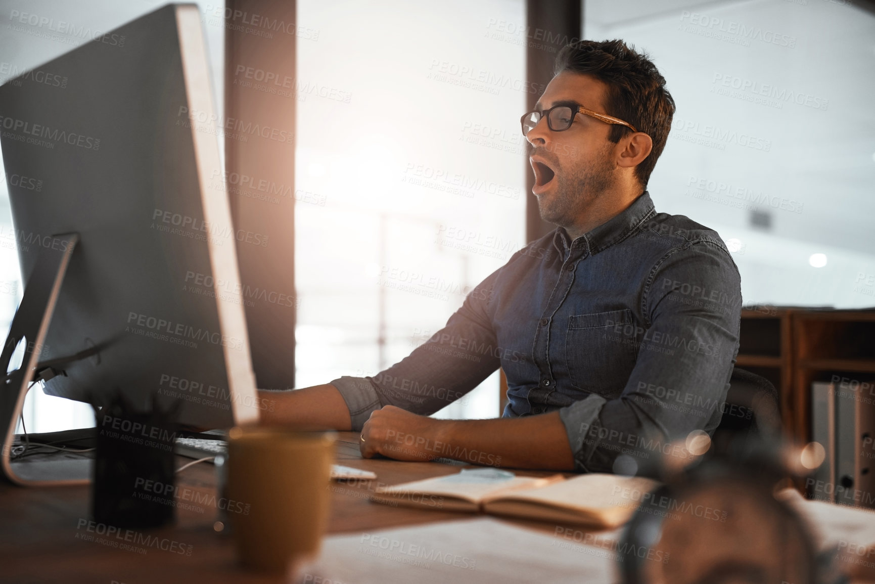 Buy stock photo Shot of a young businessman yawning while working late on a computer in an office