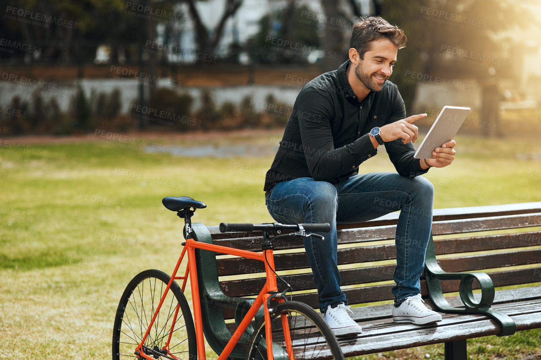 Buy stock photo Full length shot of a handsome young man using his tablet while sitting on a park bench
