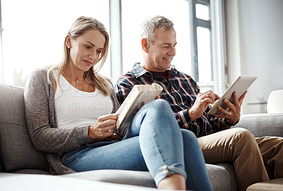 Buy stock photo Shot of a mature woman reading a book while her husband uses a digital tablet on the sofa at home