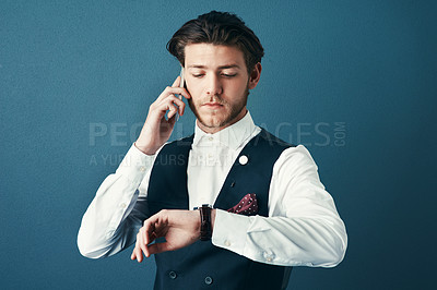 Buy stock photo Studio shot of a handsome young businessman checking the time while making a phonecall against a blue background