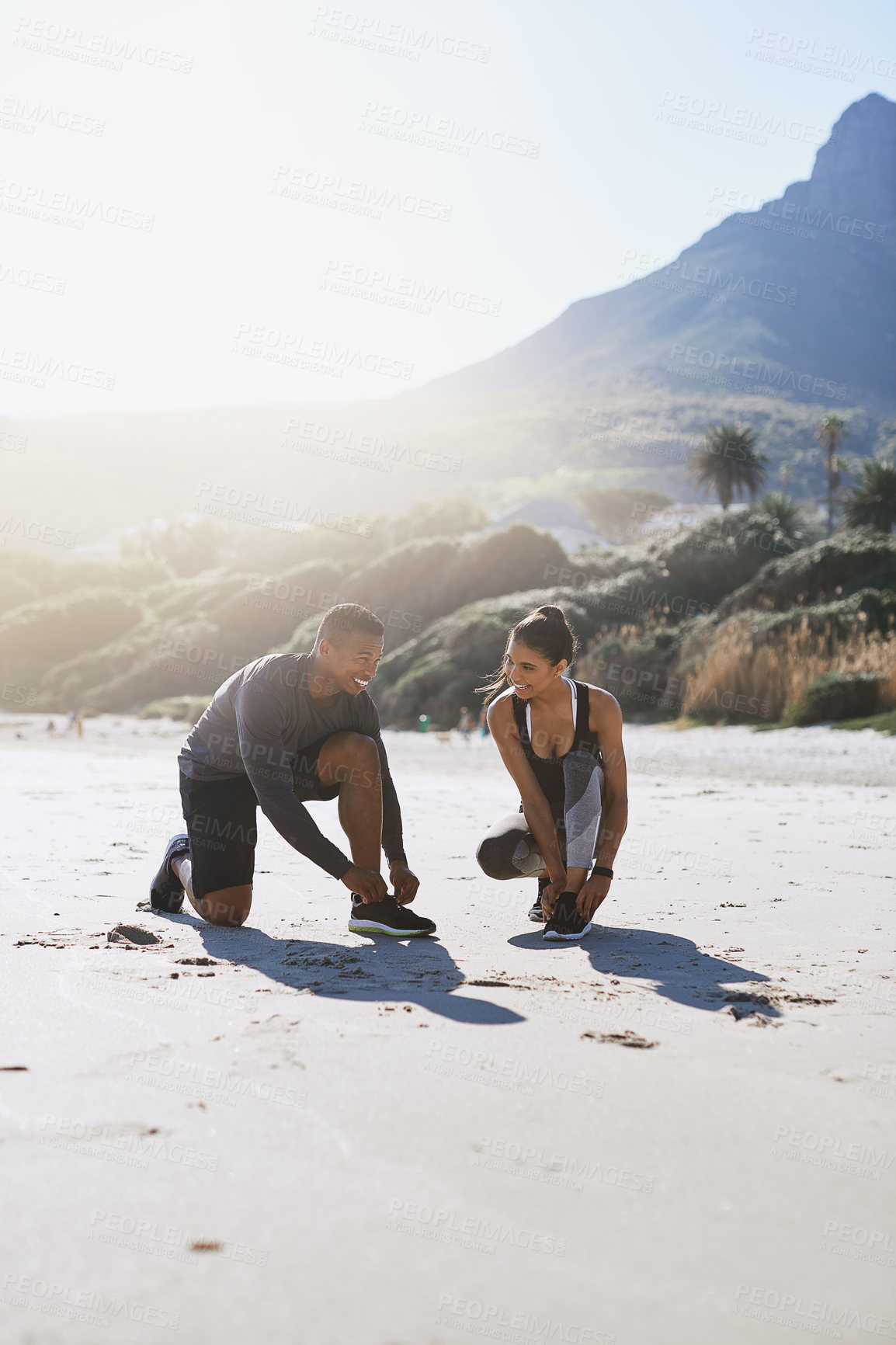 Buy stock photo Shot of a sporty young couple tying their shoelaces while out for a run along the beach