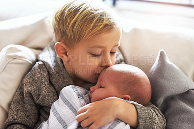 Buy stock photo Shot of a cheerful little boy holding his little infant brother and giving him a kiss on the forehead while being seated on a sofa at home during the day