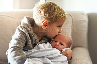 Buy stock photo Shot of a cheerful little boy holding his little infant brother and giving him a kiss on the forehead while being seated on a sofa at home during the day