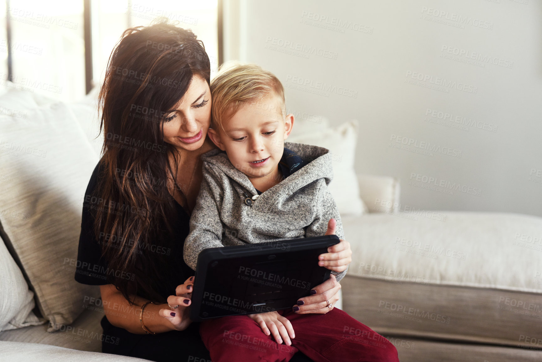 Buy stock photo Shot of a carefree young woman browsing on a digital tablet with her little boy while being seated on a sofa at home during the day