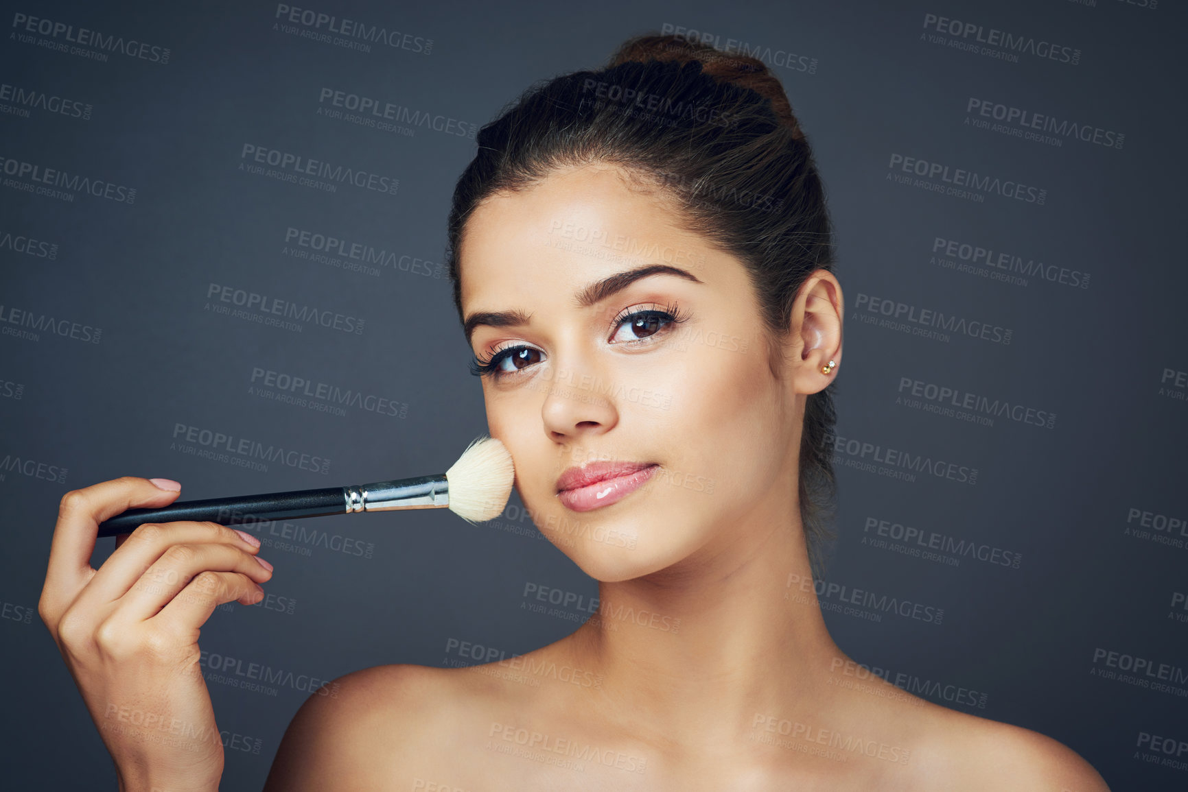 Buy stock photo Studio shot of a beautiful young woman applying blush on her cheeks while posing against a blue background