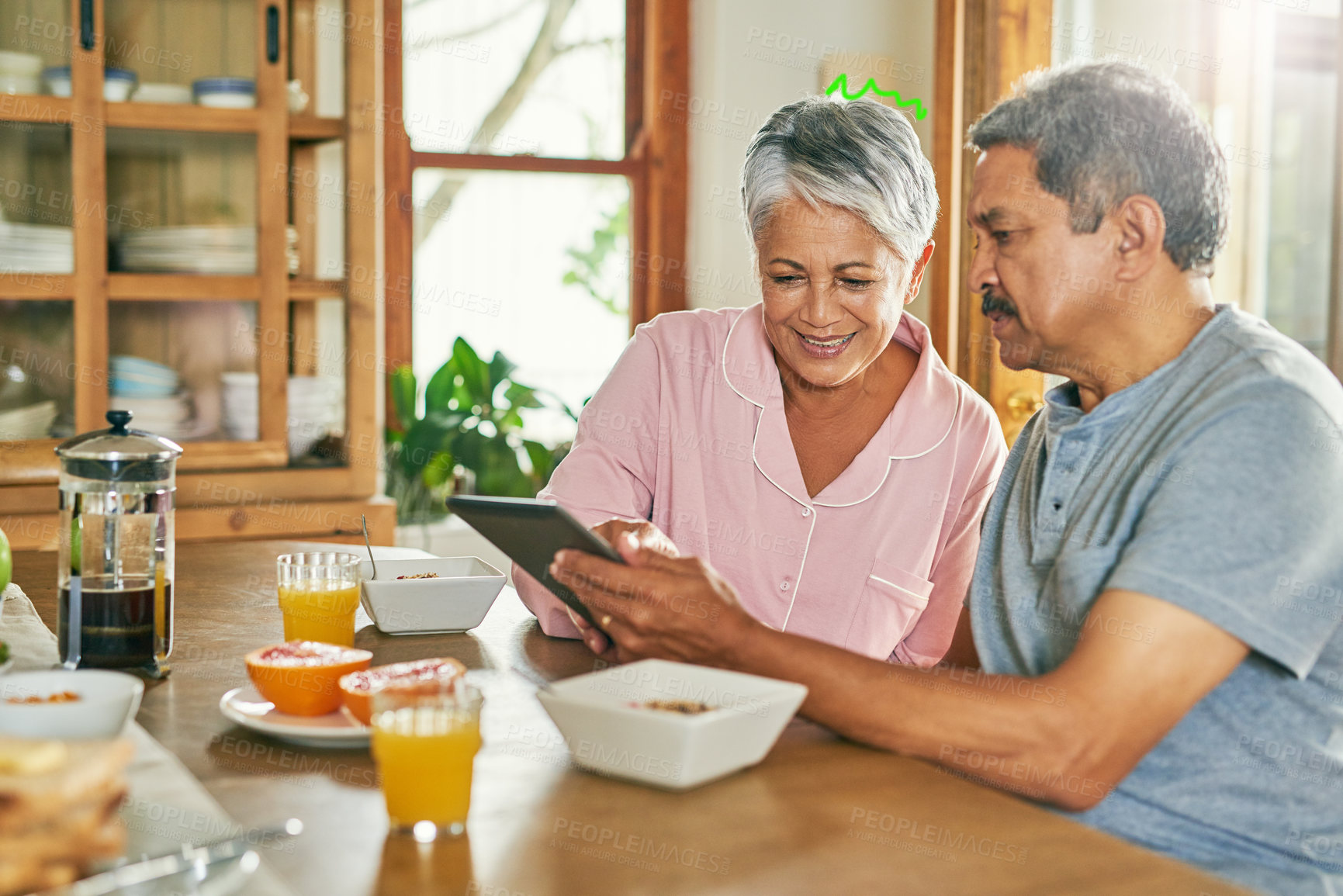 Buy stock photo Shot of a cheerful elderly couple browsing on a digital tablet together while having breakfast around a table at home