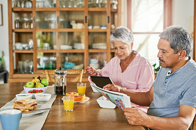 Buy stock photo Shot of a carefree elderly couple having breakfast together while reading the paper and browsing on a digital tablet at home
