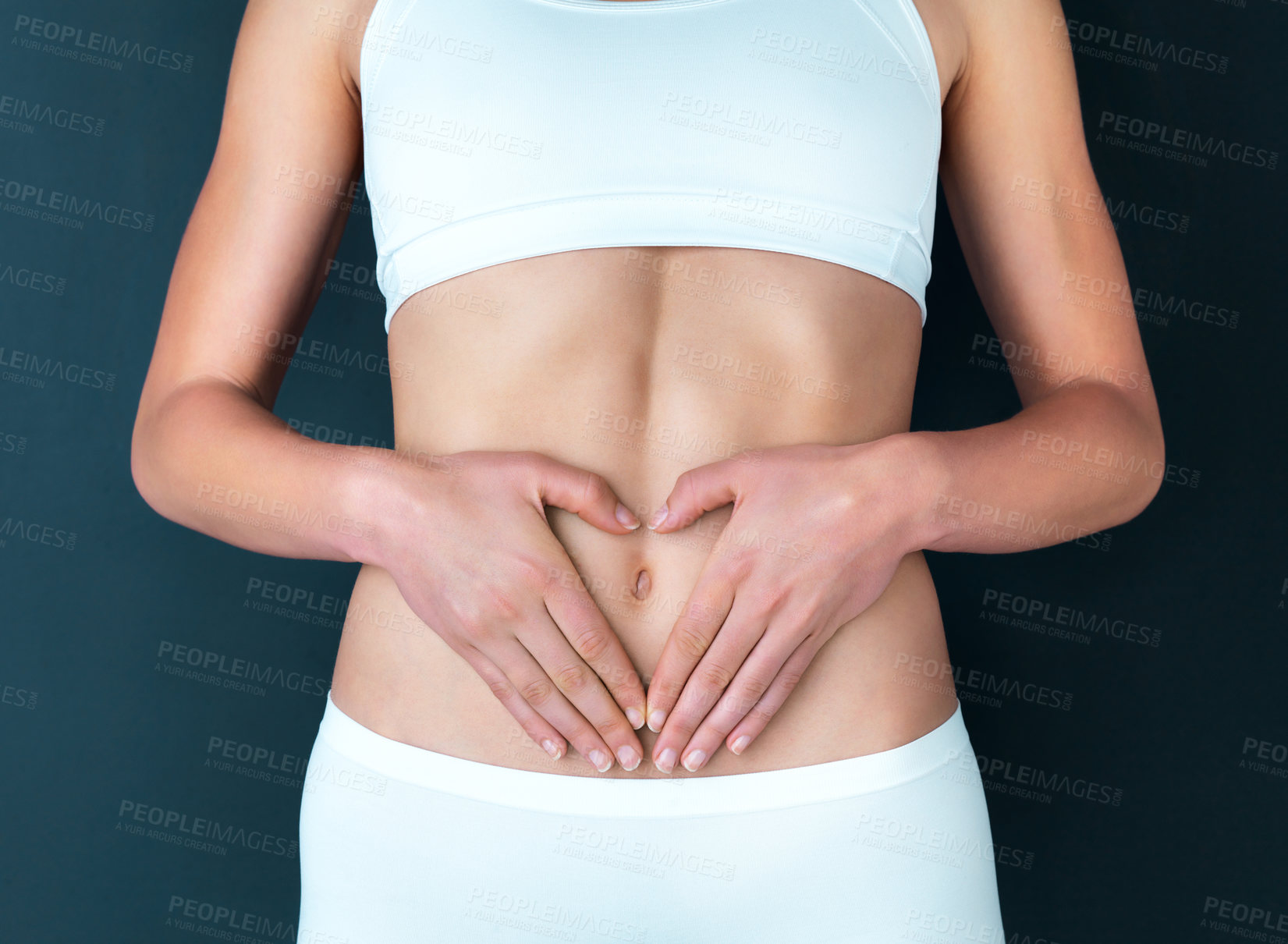 Buy stock photo Studio shot of a fit young woman making a heart shaped gesture over her stomach against a dark background