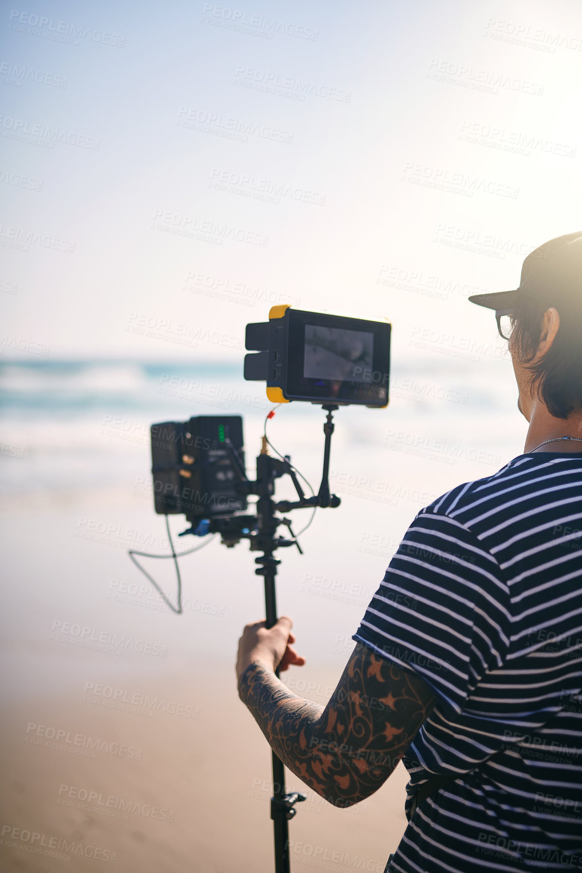 Buy stock photo Rearview shot of a focused young man shooting a scene with a state of the art video camera outside on a beach during the day