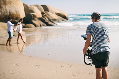 Buy stock photo Rearview shot of a focused young man shooting a scene with a state of the art video camera outside on a beach during the day