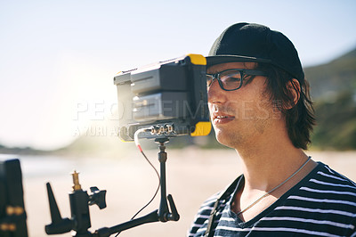 Buy stock photo Shot of a focused young man shooting a scene with a state of the art video camera outside on a beach during the day