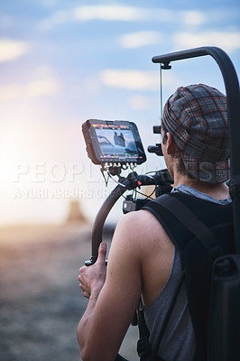 Buy stock photo Over the shoulder shot of a focused young man shooting a scene with a state of the art video camera outside on a beach during the day
