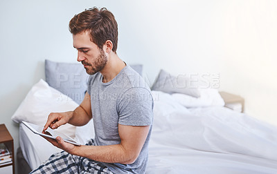 Buy stock photo Shot of a handsome young man using a tablet while sitting on a bed at home