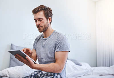 Buy stock photo Shot of a handsome young man using a tablet while sitting on a bed at home