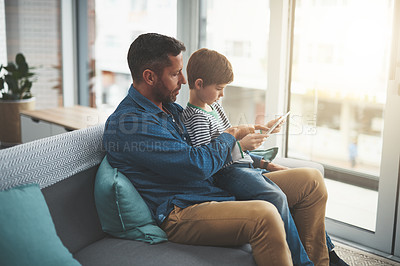 Buy stock photo Shot of a cheerful little boy browsing on a digital tablet while being seated on his father's lap at home during the day