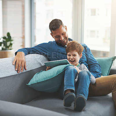 Buy stock photo Shot of a cheerful little boy browsing on a digital tablet while lying on his father's lap at home during the day