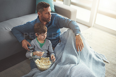 Buy stock photo Shot of a carefree young boy and his father watching a movie together while being seated on the floor and eating popcorn at home during the day