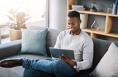 Buy stock photo Shot of a handsome young man using his digital tablet while sitting on a sofa at home