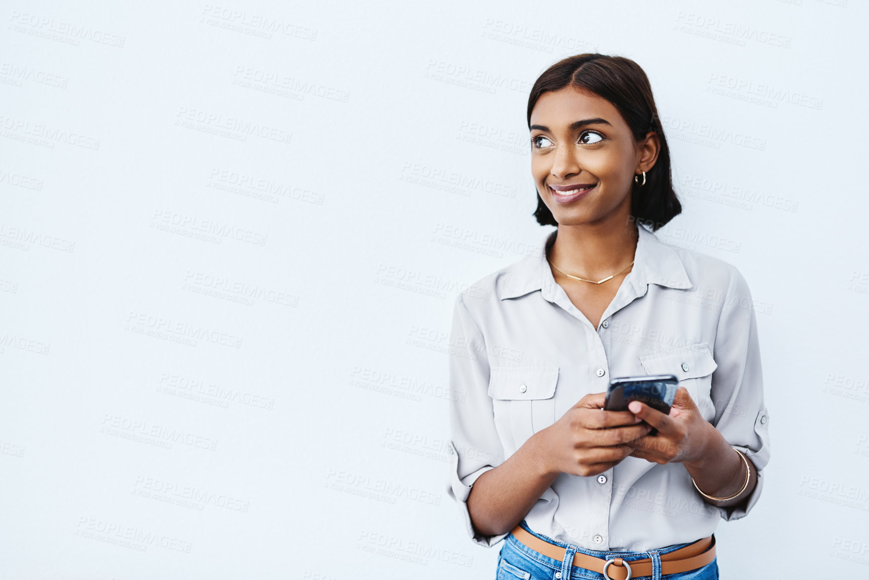 Buy stock photo Social media, young Indian woman on smartphone and thinking in studio background. Smiling or idea, phone for communication or networking and happy female person standing with cellphone in backdrop