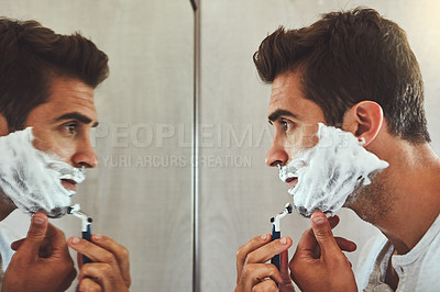 Buy stock photo Cropped shot of a handsome young man shaving his beard while looking at his reflection in the mirror at home