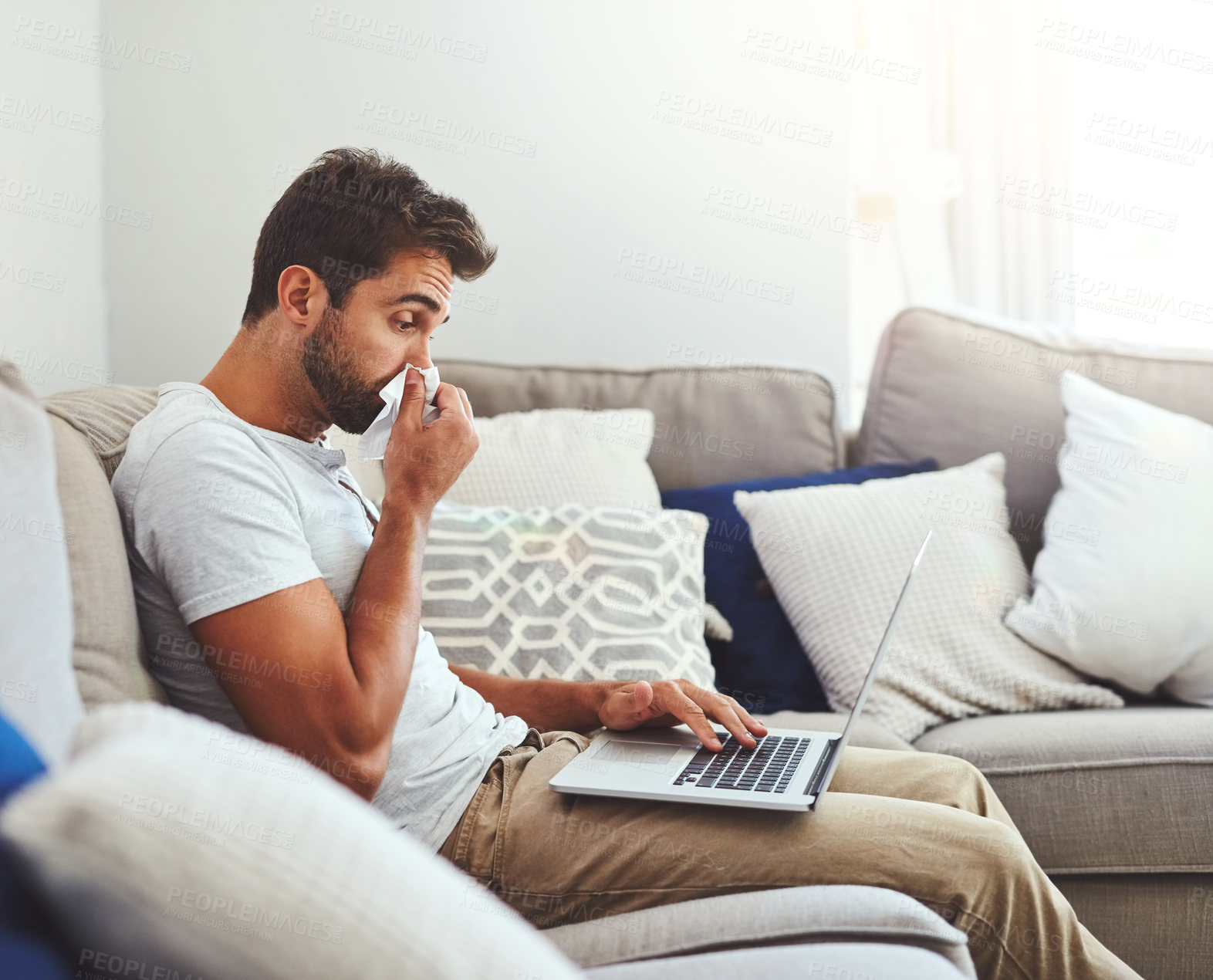 Buy stock photo Cropped shot of a handsome young man sneezing into a tissue while trying to work on his laptop at home