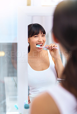 Buy stock photo Shot of a cheerful attractive young woman brushing her teeth while looking at her reflection in a mirror at home during the day