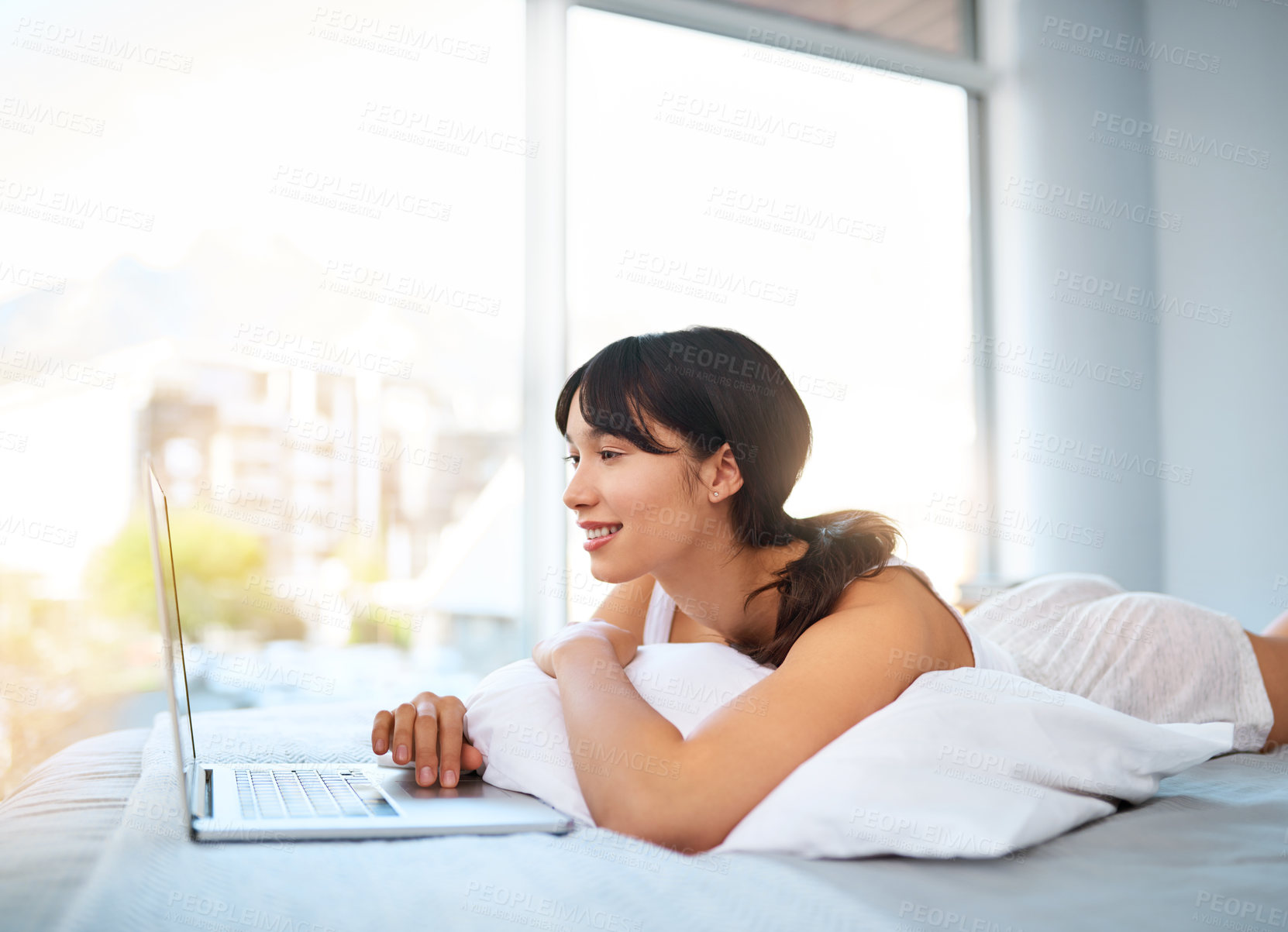 Buy stock photo Shot of a cheerful attractive young woman browsing on a laptop while lying on her bed at home during the day
