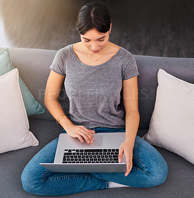 Buy stock photo Shot of a cheerful young woman browsing on her laptop while being seated on a sofa at home during the day