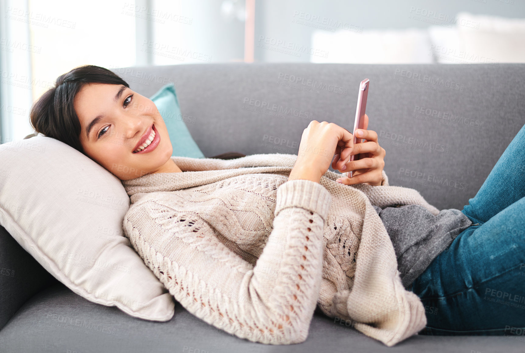 Buy stock photo Portrait of a cheerful young woman browsing on her cellphone while relaxing on a sofa at home during the day