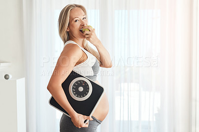 Buy stock photo Portrait of a happy pregnant woman dressed in sportswear and carrying a scale while eating an apple at home