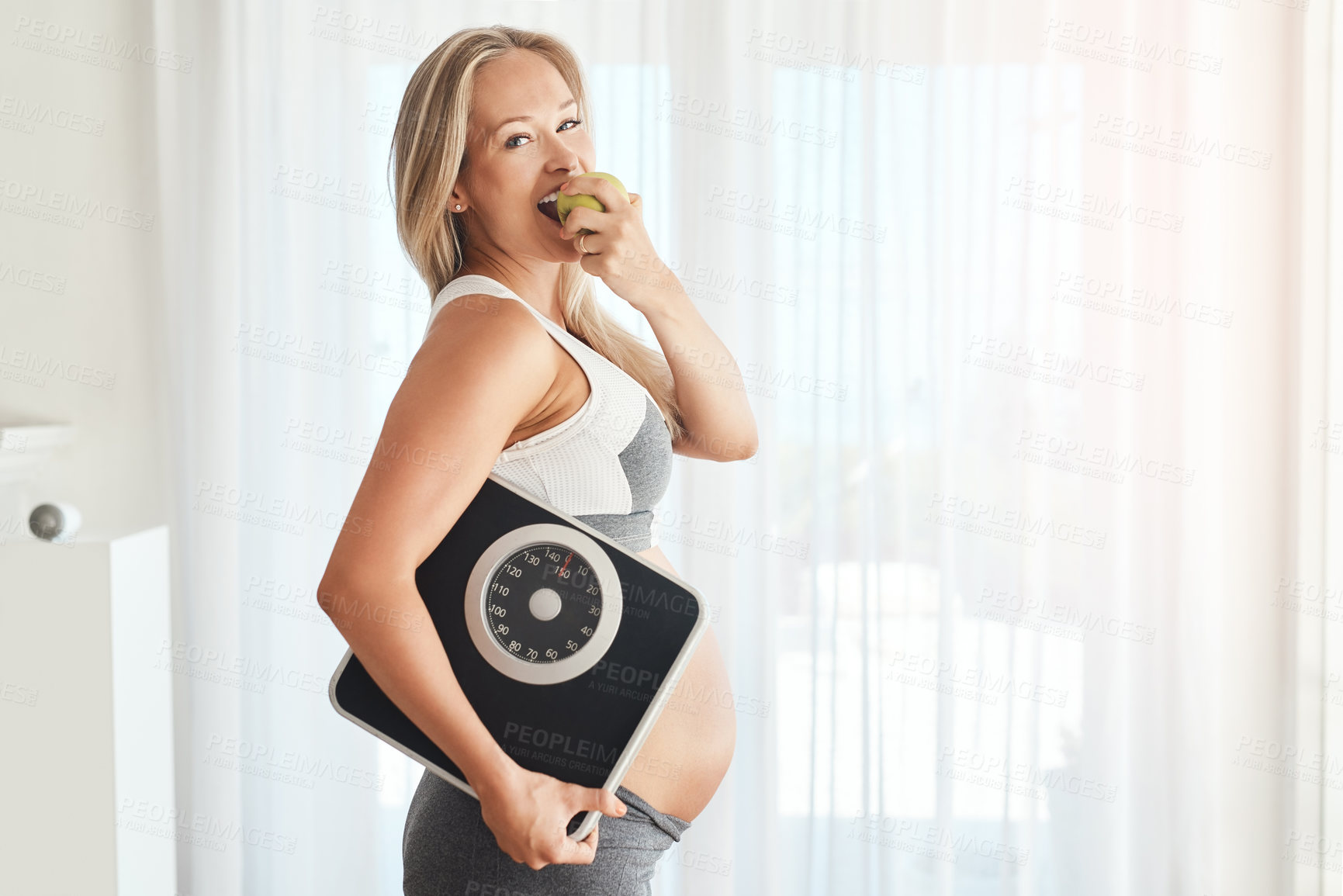 Buy stock photo Portrait of a happy pregnant woman dressed in sportswear and carrying a scale while eating an apple at home