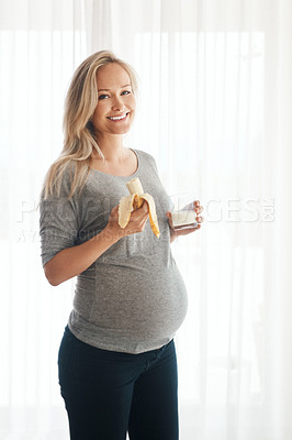 Buy stock photo Portrait of a happy pregnant woman eating a banana and drinking a glass of milk at home