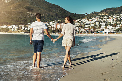 Buy stock photo Rearview shot of a carefree young couple walking hand in hand along the beach